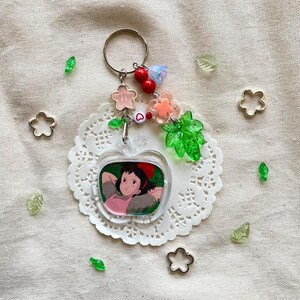 May include: A clear apple-shaped keychain featuring a cartoon character, adorned with pink and clear flower beads, red berries, and a green leaf charm. The keychain hangs from a silver ring, with decorative flower and leaf accents.
