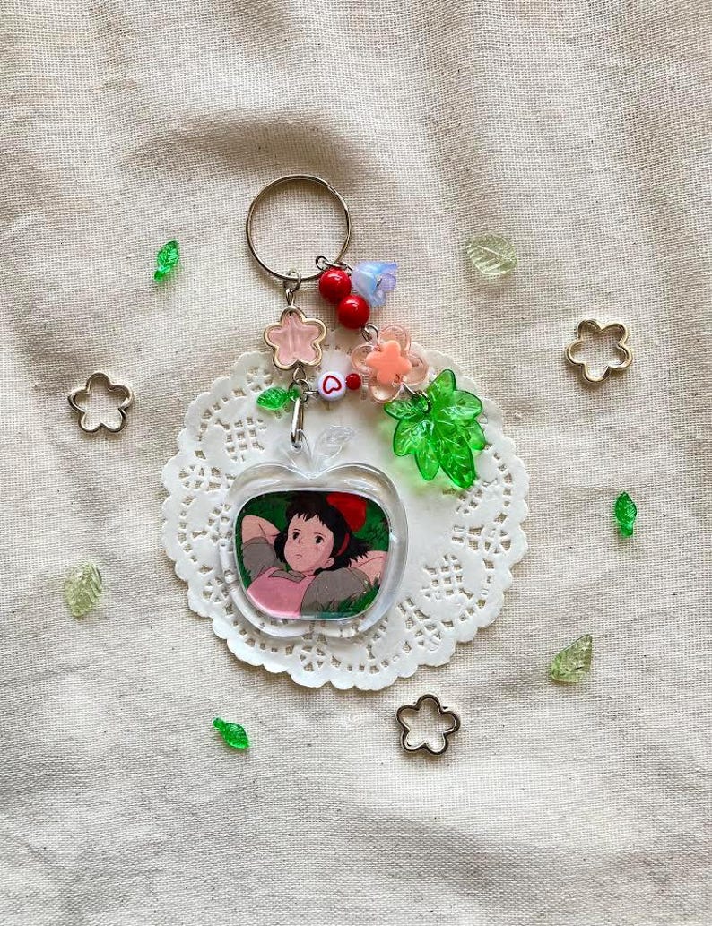 May include: A clear apple-shaped keychain featuring a cartoon character, adorned with pink and clear flower beads, red berries, and a green leaf charm. The keychain hangs from a silver ring, with decorative flower and leaf accents.