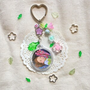 May include: A clear acrylic keychain with a heart-shaped silver ring. The keychain features a circular image of an anime character, surrounded by pink, purple, and green floral beads. Additional decorative elements include clear green leaf and flower-shaped accents.