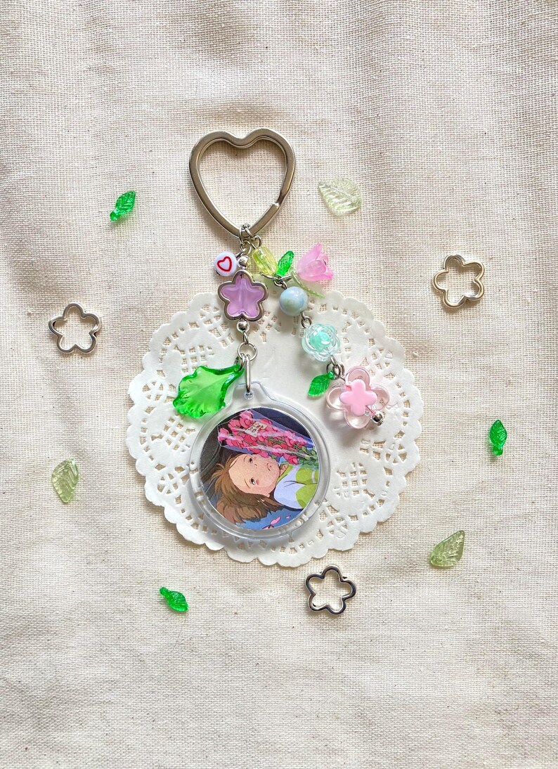 May include: A clear acrylic keychain with a heart-shaped silver ring. The keychain features a circular image of an anime character, surrounded by pink, purple, and green floral beads. Additional decorative elements include clear green leaf and flower-shaped accents.