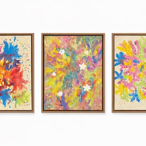 Puede incluir: Conjunto de tres pinturas abstractas enmarcadas. Cada obra presenta salpicaduras y pinceladas coloridas en tonos rosa, azul, amarillo y verde sobre un fondo beige. La pintura central incluye formas de flores blancas.