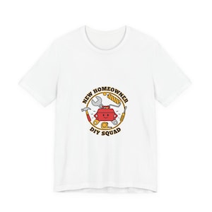 Op de afbeelding: Wit T-shirt met een cirkelvormige afbeelding met de tekst "NEW HOMEOWNER DIY SQUAD". De afbeelding bevat een gereedschapskist, moersleutel, hamer, schroevendraaier en ander gereedschap in rood, geel en zwart.