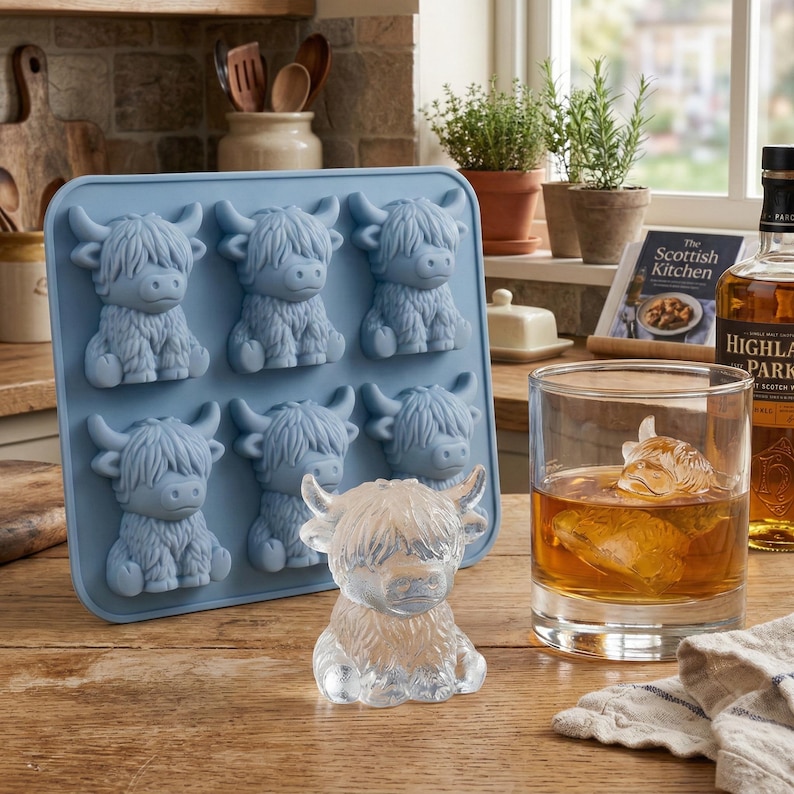 May include: A light blue silicone ice cube tray with eight Highland cow-shaped molds. A clear ice cube in the shape of a Highland cow sits next to a glass of whiskey with a Highland cow ice cube. A bottle of Highland Park scotch is in the background.