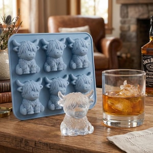 May include: A light blue silicone ice cube tray with six Highland cow molds. A clear ice cube cow sits next to a glass of amber liquid with a Highland cow ice cube. A bottle of whiskey is in the background.
