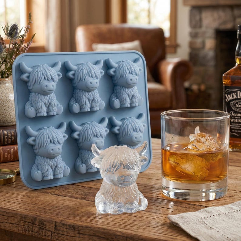 May include: A light blue silicone ice cube tray with six Highland cow molds. A clear ice cube cow sits next to a glass of amber liquid with a Highland cow ice cube. A bottle of whiskey is in the background.