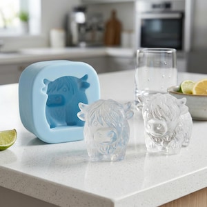 May include: A light blue silicone mold and two clear ice sculptures shaped like Highland cows sit on a white countertop. A glass and a bowl of citrus slices are in the background. The ice sculptures are detailed with shaggy fur.