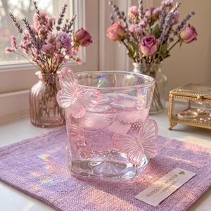 May include: A clear glass tumbler with two pink butterfly embellishments, filled with a pink liquid and ice cubes. The glass sits on a lavender cloth, with dried flowers in the background. A small tag reads "Iridescent Wonder - Handmade".