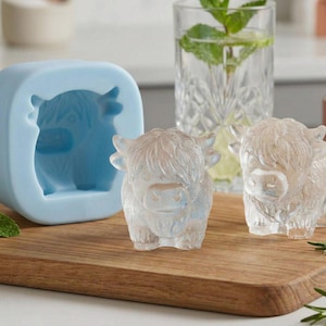May include: A light blue silicone mold and two ice cubes shaped like Highland cows sit on a wooden cutting board. The ice cubes are clear and have a detailed design. A glass of water with mint is in the background.