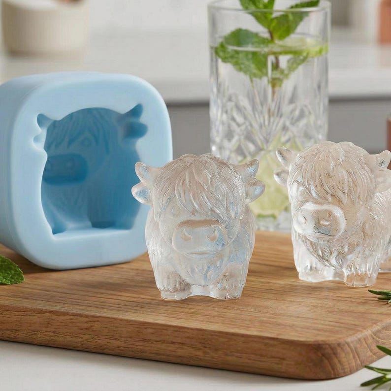 May include: A light blue silicone mold and two ice cubes shaped like Highland cows sit on a wooden cutting board. The ice cubes are clear and have a detailed design. A glass of water with mint is in the background.