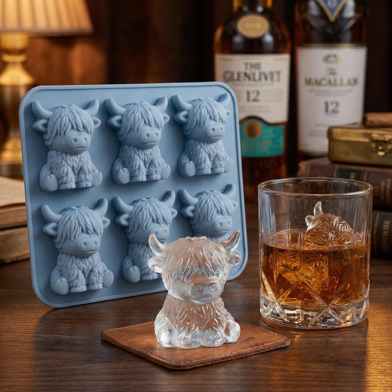 May include: A light blue silicone ice cube tray with six Highland cow shapes. A clear ice sculpture of a Highland cow sits on a brown coaster. A glass of whiskey with a Highland cow ice cube is on the right.