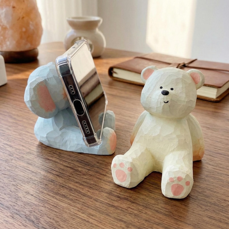 May include: A light blue bear-shaped phone holder with a phone in a clear case. A white bear-shaped phone holder is also visible. Both are made of carved wood and sit on a wooden surface.