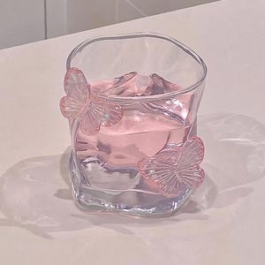 May include: A clear, square-shaped glass filled with pink liquid. Two pink butterfly embellishments adorn the glass. The glass sits on a white surface, creating a soft, pastel aesthetic. The glass is ideal for serving beverages.