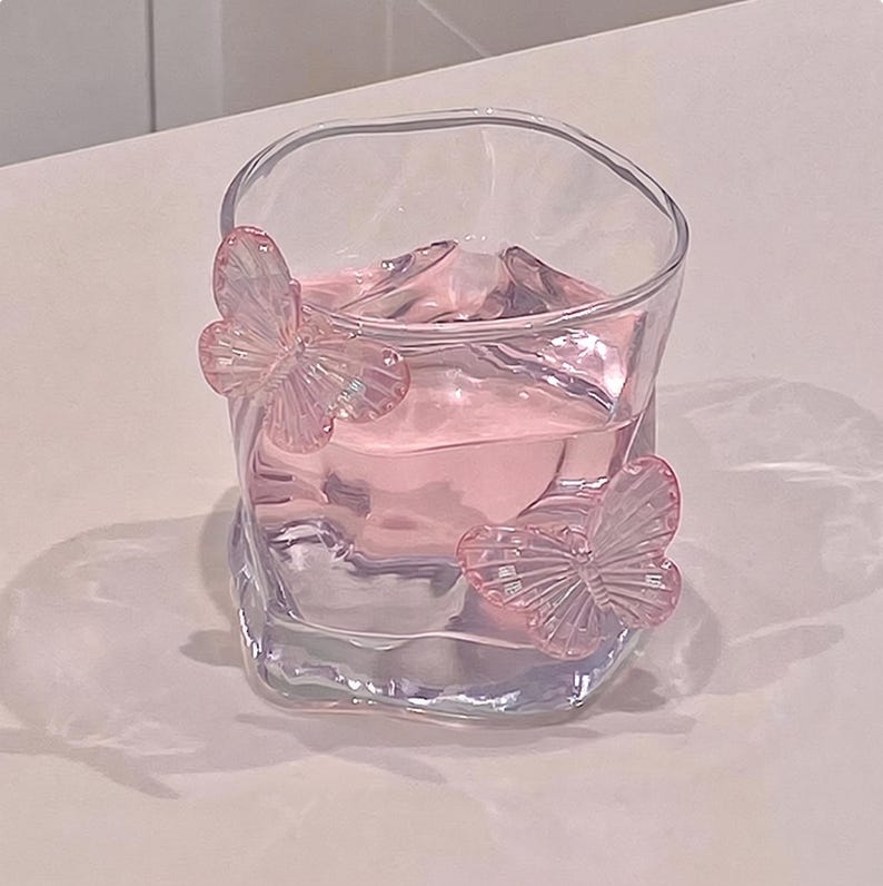 May include: A clear, square-shaped glass filled with pink liquid. Two pink butterfly embellishments adorn the glass. The glass sits on a white surface, creating a soft, pastel aesthetic. The glass is ideal for serving beverages.
