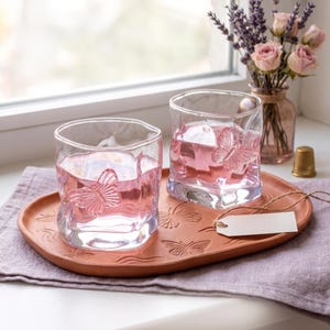 May include: Two clear, square-shaped glasses with butterfly designs, filled with pink liquid, sit on a terracotta tray. The tray also features butterfly patterns. A small vase of pink roses and lavender is in the background.