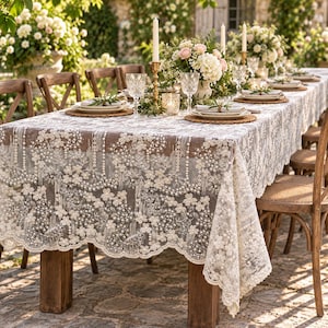 May include: A white lace tablecloth featuring an intricate floral pattern with embroidered details and small pearl accents. The fabric has a scalloped edge and drapes over a rectangular table set with plates, glassware, and floral centerpieces.