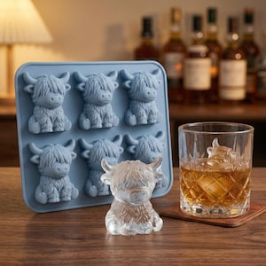 Może przedstawiać: Niebieska silikonowa forma do lodu z sześcioma formami krowy szkockiej. Przezroczysta kostka lodu w kształcie krowy szkockiej znajduje się obok szklanki bursztynowego płynu z kostką lodu w kształcie krowy szkockiej. Taca i kostki lodu znajdują się na drewnianej powierzchni.