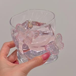 May include: A clear glass with a pink liquid inside, adorned with two pink butterfly embellishments. The glass has an irregular shape and is held by a hand with pink nail polish. The butterflies appear to be made of a translucent material.