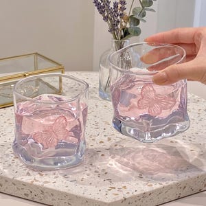 May include: Two clear, square-shaped glasses filled with pink liquid, each featuring a pink butterfly design. The glasses sit on a speckled surface, with a small vase of lavender in the background.