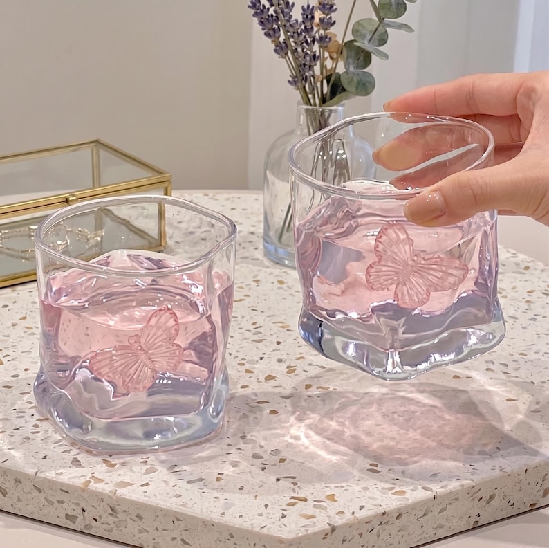 May include: Two clear, square-shaped glasses filled with pink liquid, each featuring a pink butterfly design. The glasses sit on a speckled surface, with a small vase of lavender in the background.