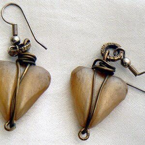 May include: A pair of handmade earrings featuring heart-shaped pendants. The pendants are a light brown color, wrapped with bronze-colored wire. The earrings have a hook closure and are displayed on a white surface.