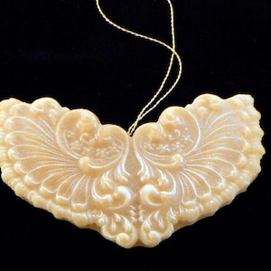 May include: A cream-colored, heart-shaped ornament with an ornate, symmetrical design. The ornament features a detailed, swirling pattern and is suspended by a gold string against a black background. The design is reminiscent of vintage decor.