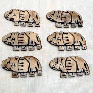 May include: Six carved, tan-colored hippo-shaped buttons with the letter "A" etched on their sides. The buttons have dark brown etched details and are arranged on a light-colored surface. Each button is approximately 1.5 inches long.