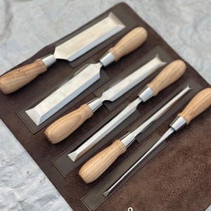 May include: A set of four woodworking chisels with polished metal blades and light-colored wooden handles, arranged diagonally in a dark brown leather tool roll. Each chisel features a silver-toned metal ferrule connecting the blade to the handle.