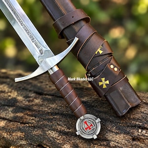May include: A steel sword with a brown leather-wrapped handle and a pommel featuring a red cross emblem. The blade is engraved with the text NON NOBIS DOMINE. It includes a brown leather scabbard with gold cross accents and leather straps.
