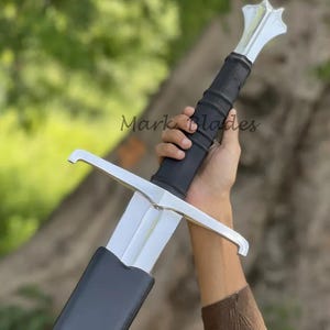 May include: A silver-colored metal sword featuring a long, straight blade, a black leather-wrapped grip, and a polished crossguard. The pommel is shaped like a star, and the blade is partially encased in a black scabbard. The text Mark Blades is visible.