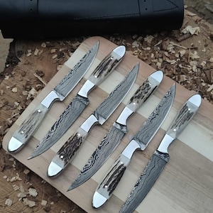 May include: A set of six fixed-blade knives with Damascus steel blades featuring wavy patterns and textured antler handles with polished metal bolsters and pommels. The knives are arranged on a light-colored wooden board near a black leather roll.
