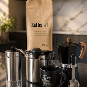Könnte beinhalten: Ein Kaffee-Set mit einer Tüte Kaffeebohnen "Kaffee art", einer Edelstahl-Kaffeemühle, einem Kaffeebehälter, einer schwarzen Tasse mit Text und einem schwarzen Espressokocher. Die Kaffeetüte trägt die Aufschrift "100% Arabica Röstkaffee" und enthält 250g Bohnen.