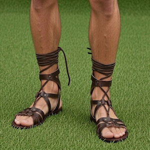 Puede incluir: Sandalias de cuero marrón con diseño de gladiador. Las sandalias tienen múltiples correas que se envuelven alrededor del tobillo y la parte inferior de la pierna. Están sobre una superficie verde.