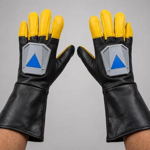 Peut inclure: Une paire de gants de style gantelet noirs et jaunes avec une longue manchette. Chaque gant présente un emblème gris en forme de bouclier sur le dos de la main, contenant un triangle bleu centré pointant vers le haut vers les doigts.