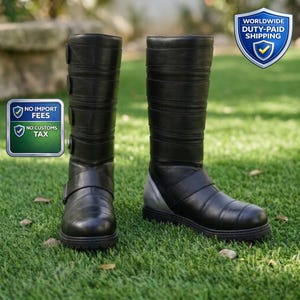 May include: A pair of black leather boots with multiple horizontal bands and silver accents. The boots have a buckle closure and are knee-high. The boots are displayed on a bed of green grass.