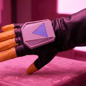 May include: Black leather glove with yellow fingertips and a gray rectangular device featuring a blue triangle design. The glove rests on a pink surface, evoking a futuristic or sci-fi aesthetic.