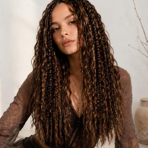 May include: A long, wavy brown wig with thin, braided sections throughout. The hair features a multi-tonal brown color palette with lighter highlights, creating a textured, voluminous appearance that falls past the shoulders in loose, crimped waves.