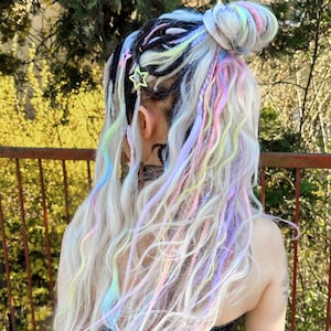 Può includere: Acconciatura con chignon e lunghe trecce ondulate. Le trecce sono un mix di nero, bianco e colori pastello, tra cui rosa, giallo e blu. I capelli sono impreziositi da fermagli a forma di stella. La persona indossa un corsetto nero e pantaloni marroni.