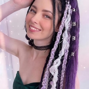 Puede incluir: Extensiones de cabello trenzadas en tonos morado, lavanda y blanco. Las trenzas están adornadas con detalles metálicos decorativos. La persona lleva un top de terciopelo verde oscuro y un choker de encaje negro.