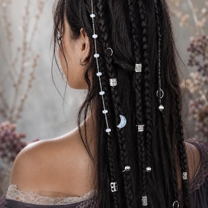 Puede incluir: Cabello oscuro peinado en múltiples trenzas adornadas con anillos, cuentas y una delicada cadena de metal plateado con pequeñas perlas blancas y un dije de media luna. Los accesorios para el cabello están distribuidos por las trenzas.