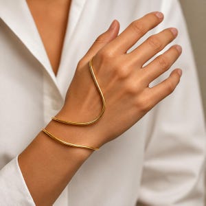 Puede incluir: Una pulsera de mano de metal dorado con un diseño de alambre liso y continuo que se envuelve alrededor de la muñeca y se extiende hacia arriba para rodear el dedo medio, creando una silueta elegante con un acabado pulido y brillante.