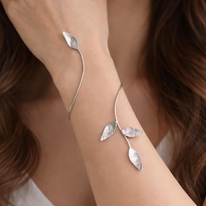 Puede incluir: Un brazalete plateado con un diseño de alambre fino y curvado que se envuelve alrededor del antebrazo. La pieza está adornada con cuatro acentos metálicos texturizados en forma de hoja, con una hoja en el extremo superior y tres abajo.