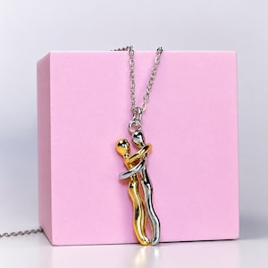 May include: A silver-toned metal chain necklace featuring a pendant of two stylized figures in an embrace. One figure is finished in a polished gold tone, while the other is finished in a polished silver tone, creating a two-tone metallic effect.