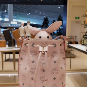 Puede incluir: Un bolso cubo rosa con cierre de cordón y el patrón del logotipo de MCM. Una cabeza de conejo de peluche con orejas largas asoma de la bolsa. El bolso tiene un ribete rosa claro y herrajes plateados.