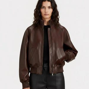 Puede incluir: Chaqueta bomber de cuero marrón con cuello, puños y dobladillo de canalé. La chaqueta tiene cierre de cremallera y dos bolsillos laterales. La modelo lleva un top negro y pantalones negros.