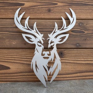 Pode incluir: Uma peça de arte de parede em metal de cor prateada com o formato da cabeça e da galhada de um veado. O design apresenta recortes e linhas angulares nítidas que formam os traços faciais e a galhada ramificada do animal num padrão simétrico.