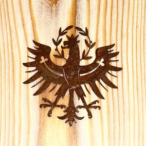 Könnte beinhalten: Ein rostbraunes Metalladler-Emblem mit ausgebreiteten Flügeln und einem Blätterkranz. Der Adler ist auf einem hellen Holzgrund mit vertikaler Maserung zentriert. Das Emblem hat komplizierte Details und ein rustikales Aussehen.