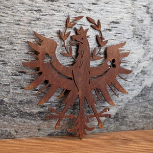 Könnte beinhalten: Eine flache, rostfarbene Wanddekoration aus Metall in Form eines stilisierten Adlers mit einer Krone auf dem Kopf und belaubten Zweigen, die sich über den Flügeln wölben. Der Adler weist filigrane Ausschnitte in den Flügeln und einen spitzen Schwanz auf.