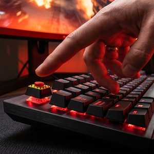 May include: A close-up of a black and gray mechanical keyboard with red backlighting. A finger is pressing a large, red button with a yellow and black hazard stripe. The keyboard is on a dark surface.