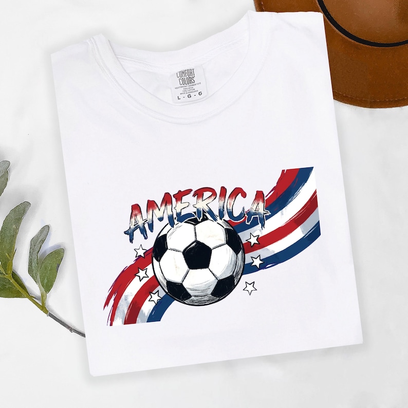May include: A white t-shirt featuring a graphic print of a black and white soccer ball centered over red, white, and blue brushstroke stripes. The word AMERICA is printed in a distressed blue font above the ball, surrounded by small white stars.