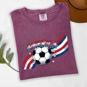May include: A maroon short-sleeved t-shirt featuring a graphic of a soccer ball centered over red, white, and blue brushstroke stripes. The word AMERICA is printed in a distressed blue font above the ball, surrounded by small white stars.